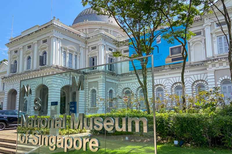 The exterior of the National Museum of Singapore with a glass sign in the foreground and a historic white building in the background—a must-see for those seeking things to do in Singapore on a budget.