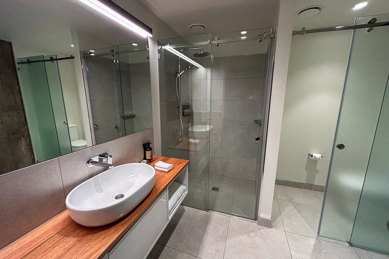Things to Do in Launceston 28 Modern Launceston bathroom with a large mirror, vessel sink on a wooden worktop, toiletries, towel, glass-enclosed shower, and grey tile flooring and walls. Toilet is visible through another glass door.