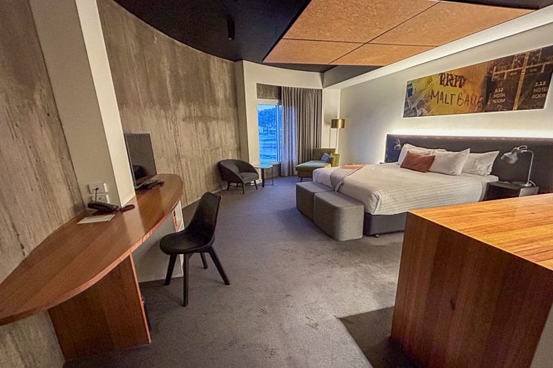 Things to Do in Launceston 27 A modern Launceston hotel room with a large bed, desk, chair, TV, a seating area near the window, and wall art above the bed. The room features neutral tones with a stylish mix of wood and concrete finishes.