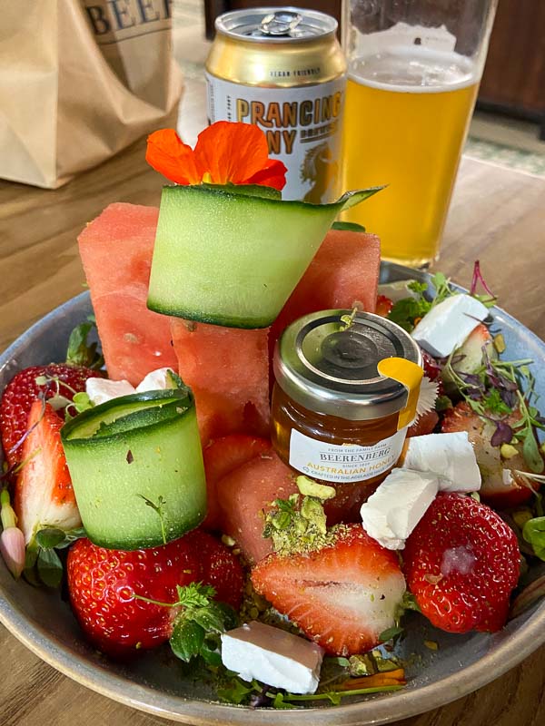 A bowl with strawberries, watermelon, cucumbers, feta, and a small jar of honey. A can and half-full glass of beer are in the background.