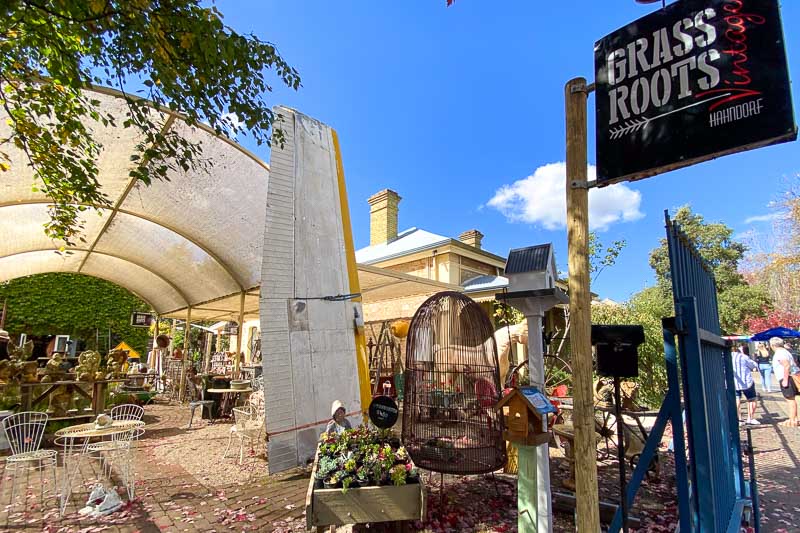 Outdoor café with seating under a canopy, surrounded by greenery and rustic decor. A sign reads "Grass Roots Hahndorf" under a clear blue sky.