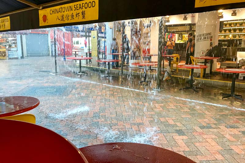 Heavy rain pours from a roof onto outdoor seating at a Chinatown seafood restaurant, creating puddles on the ground.