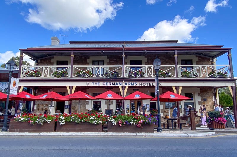 Historic two-story stone hotel with balcony, red umbrellas, and outdoor seating adorned with colorful flowers, under a blue sky with clouds.