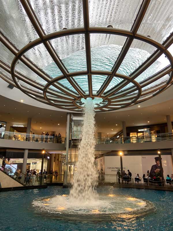 Indoor waterfall feature in a shopping mall, with water cascading from a circular metal structure into a pool below, surrounded by shops and seated visitors.