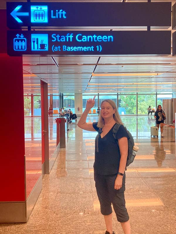 What I Packed - 8 weeks, Hot & Cold 4 Person in black clothes and backpack pointing to an overhead sign for a lift and staff canteen in an indoor setting.