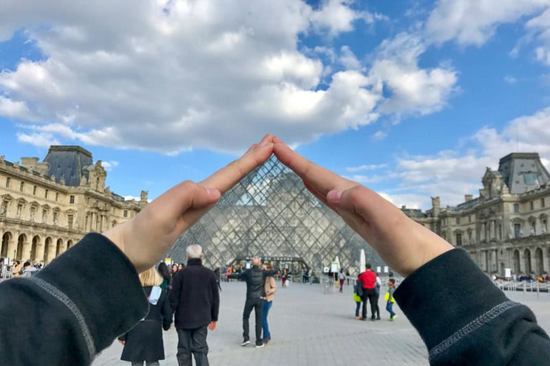 Travel Diaries - Paris 2 Hands framing the glass pyramid at the Louvre Museum entrance, with people walking around under a partly cloudy sky.