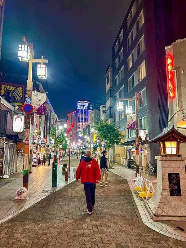 Tokyo Free Walking Tours: 3 Tours I Tried and Recommend 8 A person in a red jacket walks down a brightly lit city street at night, surrounded by buildings, signs, and a few other pedestrians.