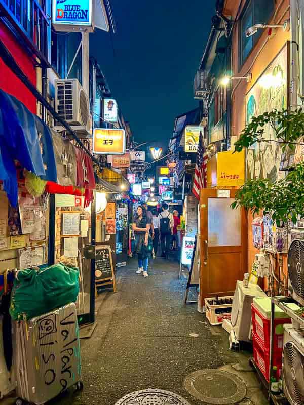 Tokyo Free Walking Tours: 3 Tours I Tried and Recommend 24 A narrow, brightly lit street lined with bars, signs, and posters at night, with people walking and various items and air conditioners along the sides.
