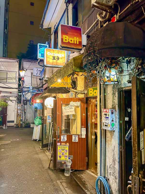 Tokyo Free Walking Tours: 3 Tours I Tried and Recommend 23 Narrow alley at night with illuminated signs, including "Bali," outside small bars and restaurants in an urban setting. String lights and other decorations are visible.