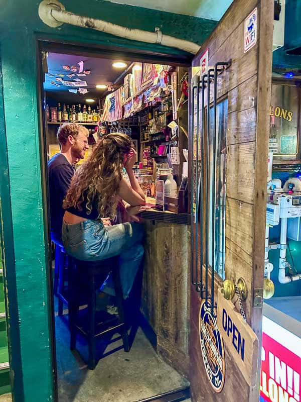 Tokyo Free Walking Tours: 3 Tours I Tried and Recommend 25 Two people sit on stools at a small bar counter inside a narrow, colorful establishment with various items and signs visible on the walls and door.
