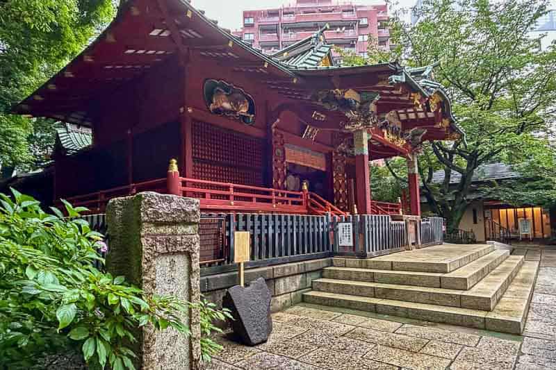 Tokyo Free Walking Tours: 3 Tours I Tried and Recommend 10 Traditional Japanese shrine with red wooden structure, ornate roof, and stone steps, surrounded by trees and modern buildings in the background.