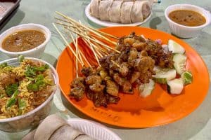 Singapore Hawker Centres You Should Try 38 Grilled meat skewers on an orange plate with sliced cucumbers, served with dipping sauces and side dishes on a table.