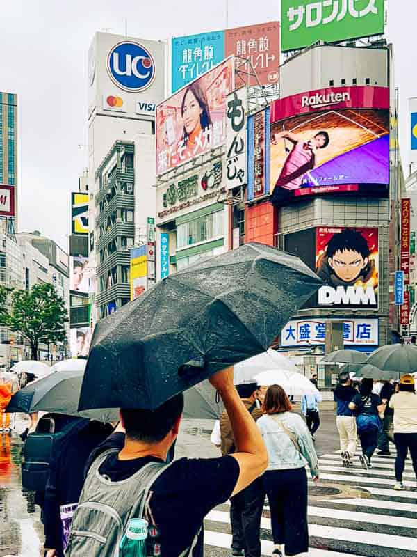 Tokyo Free Walking Tours: 3 Tours I Tried and Recommend 16 People with umbrellas cross a busy intersection in a city with tall buildings and colorful advertising billboards on a rainy day.