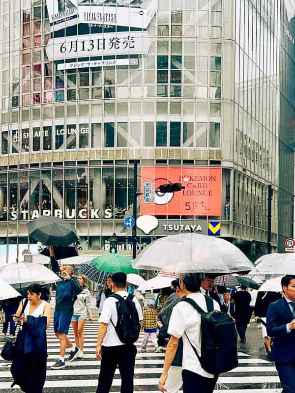 Tokyo Free Walking Tours: 3 Tours I Tried and Recommend 15 People with umbrellas cross a busy intersection in front of Starbucks and Tsutaya stores in an urban area, with digital billboards visible on the buildings.
