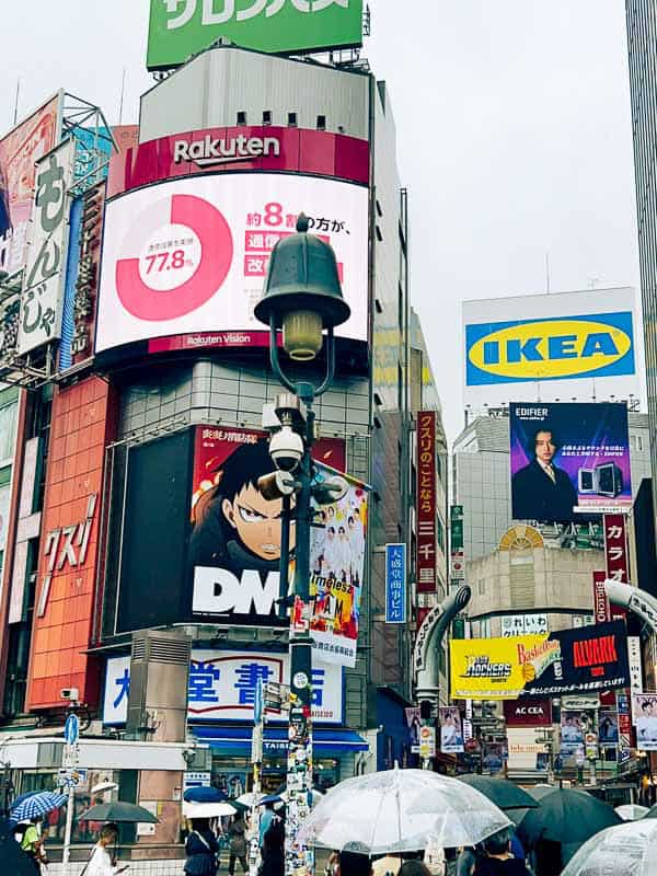 Tokyo Free Walking Tours: 3 Tours I Tried and Recommend 14 Busy city scene with colorful billboards, including ads for Rakuten, IKEA, and anime, above a crowd of people holding umbrellas on a rainy day.