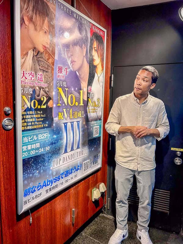Tokyo Free Walking Tours: 3 Tours I Tried and Recommend 22 A man stands indoors next to a wall with colorful posters featuring three people and Japanese text promoting "Top Dandy Three.