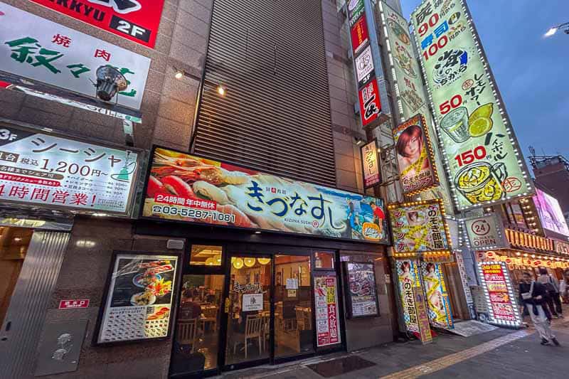 Tokyo Free Walking Tours: 3 Tours I Tried and Recommend 17 Street scene showing brightly lit restaurant signs in Japanese, with images of sushi and food, and people dining inside a sushi restaurant at dusk.