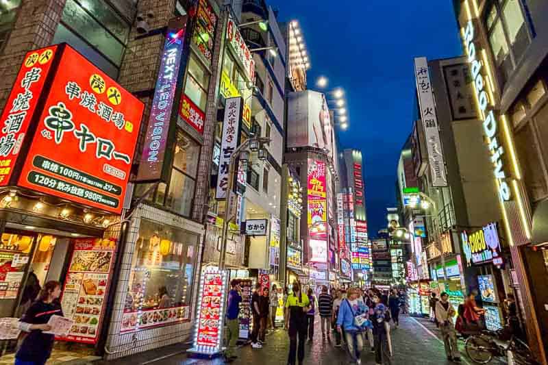 Tokyo Free Walking Tours: 3 Tours I Tried and Recommend 18 A busy city street in Japan at night with many illuminated signs, tall buildings, and people walking along the sidewalks.