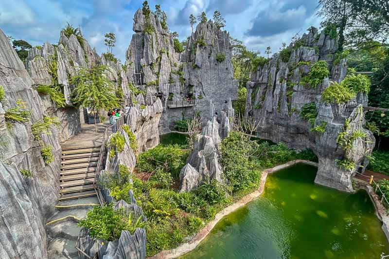 Which Mandai Wildlife Park Should You Visit? I Tried All Five 35 A winding wooden walkway passes through jagged grey limestone formations and green ponds under a partly cloudy sky, with sparse vegetation throughout the area.