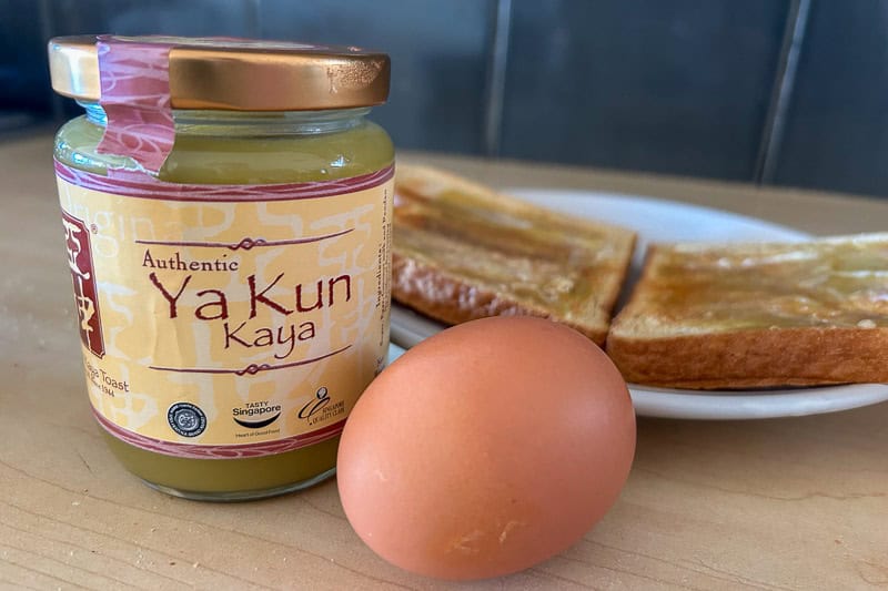 A jar of Ya Kun Kaya spread, a soft-boiled egg, and kaya toast slices on a plate sit on a wooden surface.