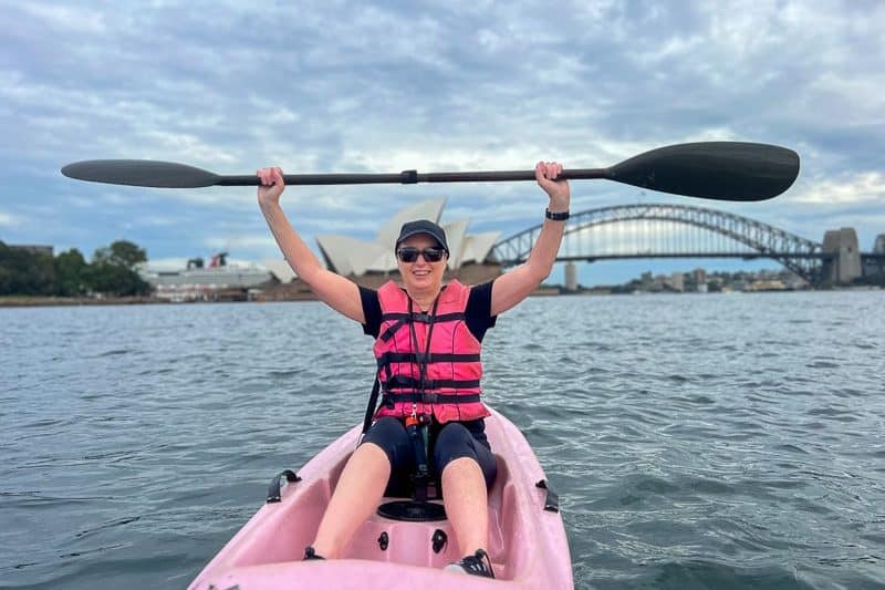 A woman in a pink life vest lifts her paddle triumphantly while kayaking on Sydney Harbour, with the iconic Sydney Opera House and Harbour Bridge in the background. The sky is overcast, adding a dramatic touch to the lively scene.