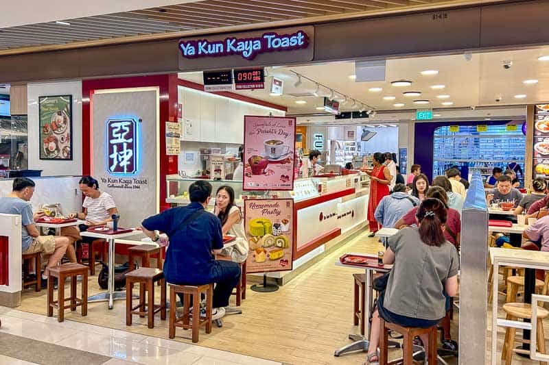 Customers seated and dining inside and outside Ya Kun Kaya Toast café in a shopping mall, with staff working behind the counter and menu signs displayed. The best place to eat kaya toast in Singapore