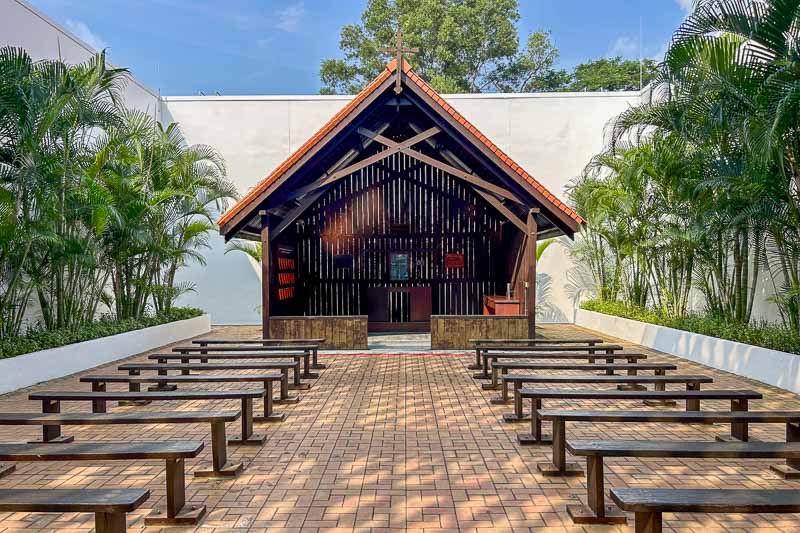 Outdoor chapel with wooden benches, brick flooring, and a small open-air structure with a peaked roof, surrounded by green plants and white walls—a peaceful stop to include in your 7 Day Singapore Itinerary.