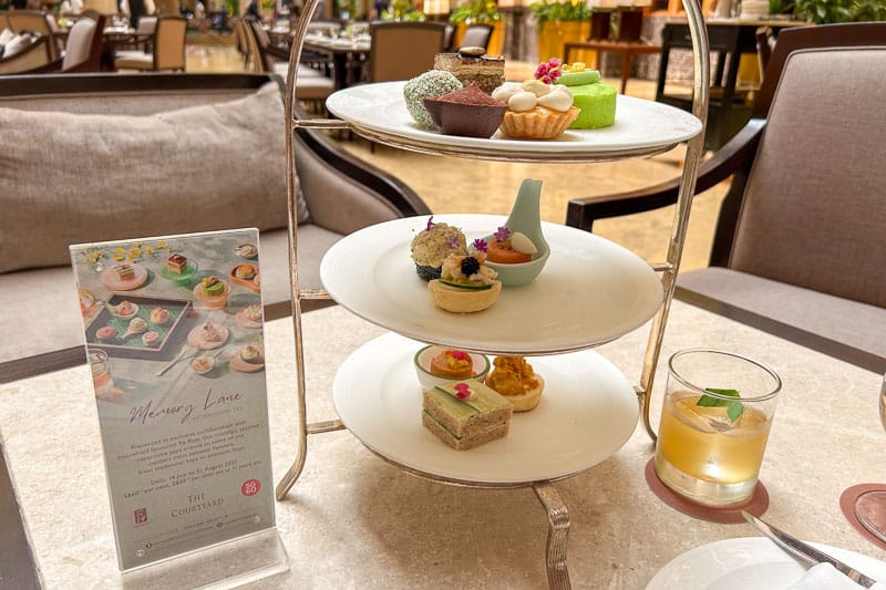 A three-tiered tray with assorted pastries and finger foods, a drink with mint, and a menu card on a marble table set the scene for a luxe lounge stop on your 7 Day Singapore Itinerary.