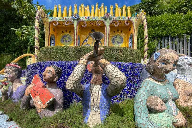 Mosaic statues of people, including one playing a trumpet, stand in front of a colorful, ornate structure with raised golden hands on top, surrounded by greenery.