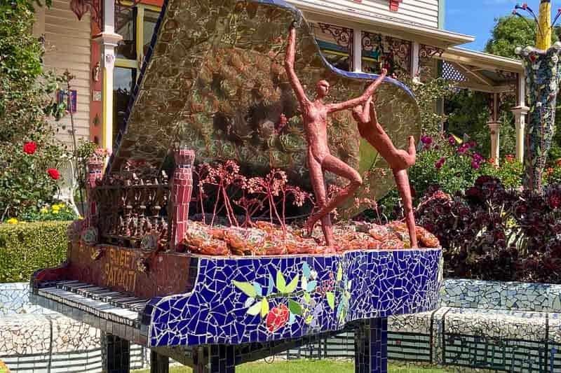 A mosaic grand piano sculpture features two abstract human figures dancing on top, surrounded by colorful patterns and garden plants.