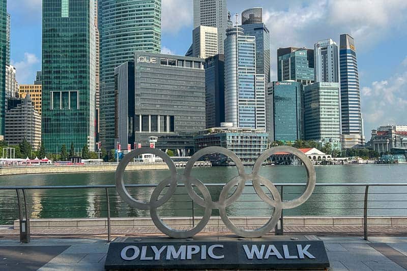 Large Olympic rings sculpture with "Olympic Walk" sign in front of a waterfront and modern skyscrapers—a must-see spot on any 2 Day Singapore Itinerary.