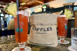 10 Day Singapore Itinerary – The Ultimate Guide 30 Two Singapore Sling cocktails garnished with pineapple sit on a table beside a burlap bag labeled "Raffles Singapore," capturing the perfect moment from a 7 Day Singapore Itinerary in an elegant restaurant setting.