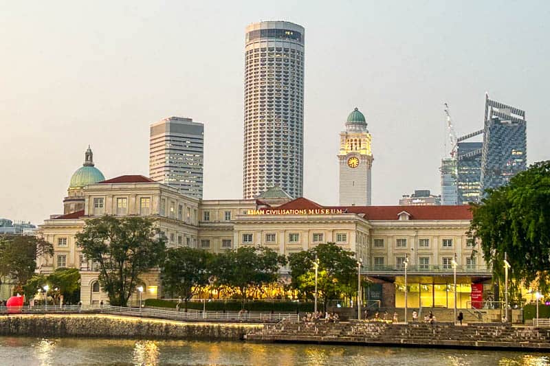 The Asian Civilisations Museum beside the Singapore River, with modern skyscrapers and a historic clock tower in the background at dusk, invites you to explore and Visit Singapore's rich heritage.