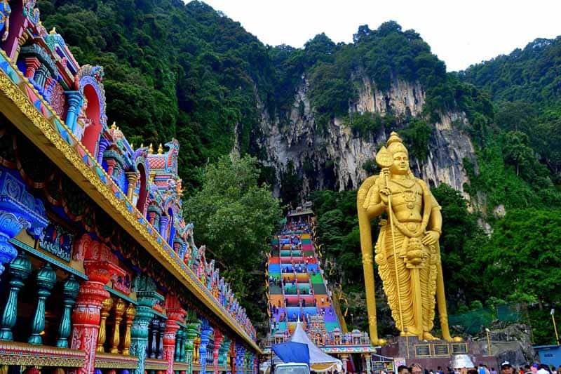 A large golden statue stands near colorful temple stairs and structures, set against a backdrop of lush green cliffs and limestone caves.
