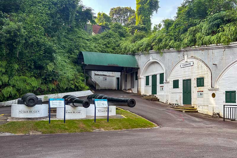 Three large artillery guns stand outside a white historical bunker with arched doorways and green shutters, surrounded by lush greenery—a unique stop to consider for your 10 Day Singapore Itinerary.