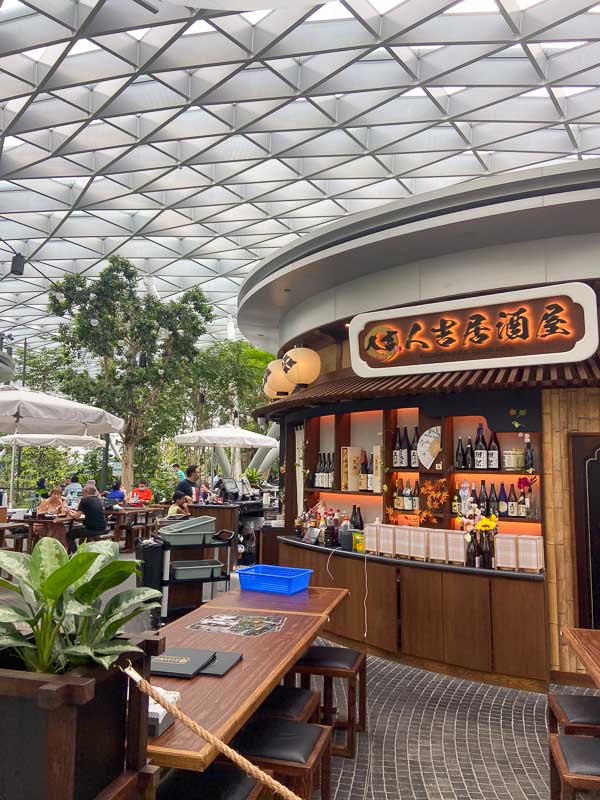 Indoor dining area with wooden tables and lush plants; this Japanese-themed restaurant at Jewel at Changi features bottles on display beneath a striking geometric glass ceiling.