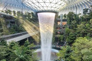 An indoor waterfall under a glass dome at Jewel at Changi, surrounded by lush greenery and walkways, with sunlight streaming through the roof.