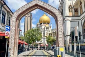 4 Day Singapore Itinerary – Best Things to Do in 4 Days 31 A stone arch frames the gold dome of Sultan Mosque in Singapore, a highlight on any 4 Day Singapore Itinerary, surrounded by shops, street signs, and pedestrians on a sunny day.