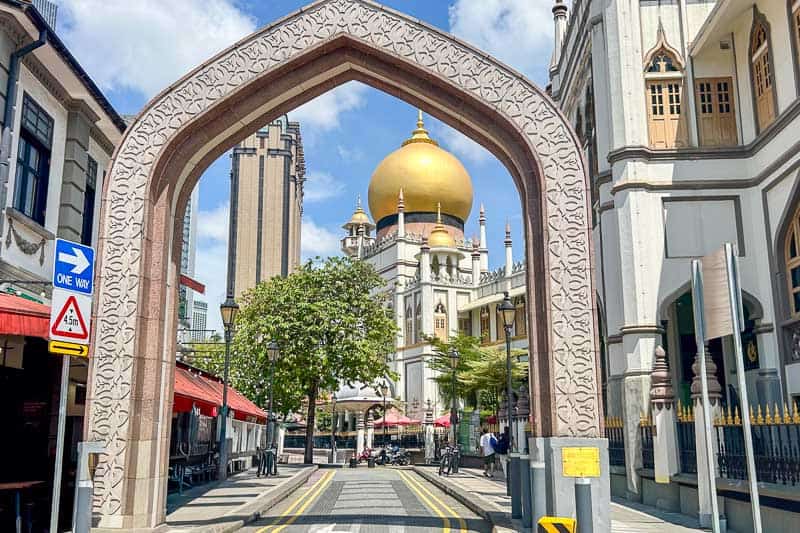 A stone arch frames the gold dome of Sultan Mosque in Singapore, a highlight on any 4 Day Singapore Itinerary, surrounded by shops, street signs, and pedestrians on a sunny day.