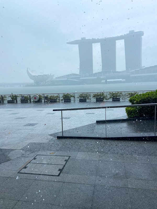 Rain falls on a waterfront promenade as the iconic Marina Bay Sands emerges through the foggy background—inviting you to visit Singapore and experience its unique skyline.