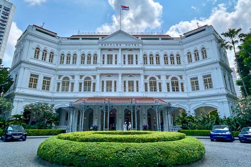 The facade of Raffles Hotel in Singapore, with white colonial architecture, a central driveway, lush greenery, and a flag on the roof, is a must-see highlight on any Singapore & Malaysia Itinerary.