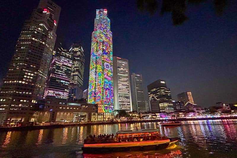 A tall skyscraper features colorful, vibrant light projections at night, surrounded by other lit buildings and a boat on the river in the foreground—an iconic scene that invites you to visit Singapore.