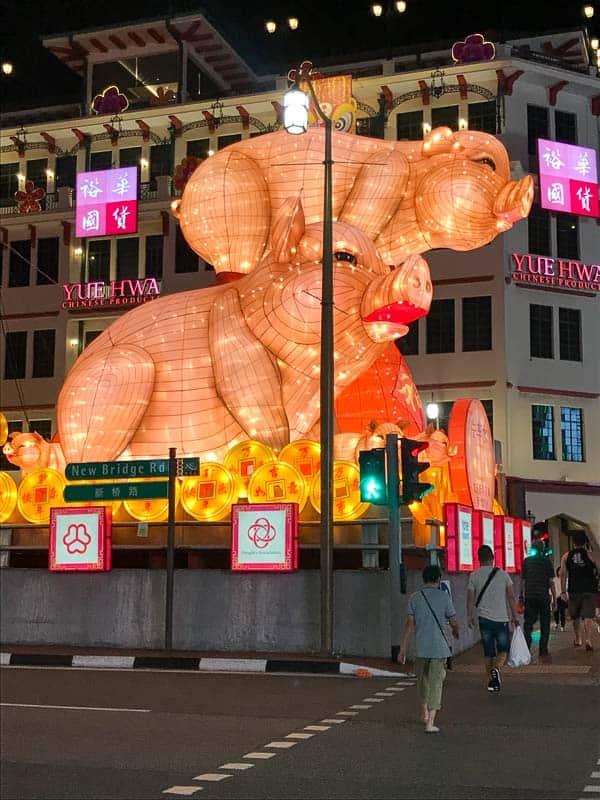 Large illuminated pig lantern display at a street intersection in front of the Yue Hwa Chinese Products building at night, with pedestrians crossing—a must-see when you Visit Singapore.