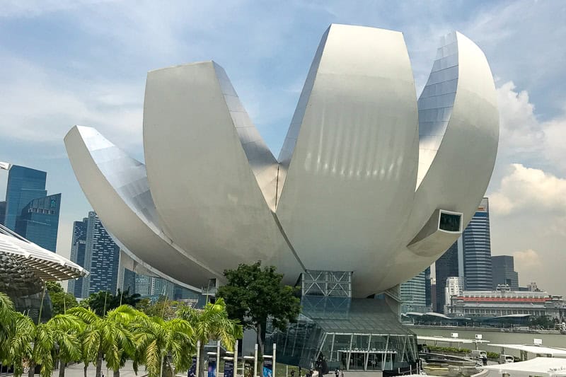 Things to Do in Singapore: A First-Timer’s Guide 6 A modern building with white petal-like structures, set against Singapore's cityscape with palm trees and soaring skyscrapers in the background.