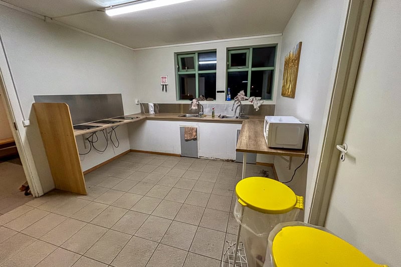 Iceland Campgrounds 3 A small, clean kitchen with two sinks, a stovetop, a microwave, and two large yellow-lidded bins. The room has tiled flooring and white walls with a window above the sinks.
