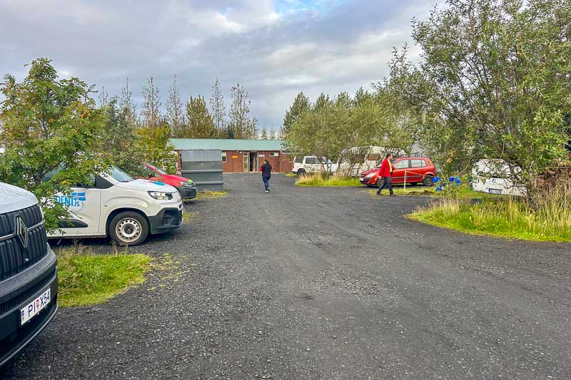 Iceland Campgrounds 2 Gravel parking area with parked cars, a person walking toward a small building, and trees surrounding the lot under a cloudy sky.