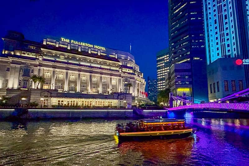 Best Areas to Stay in Singapore 6 A brightly lit boat cruises along a river at night near The Fullerton Hotel, with modern buildings and an illuminated bridge in the background.