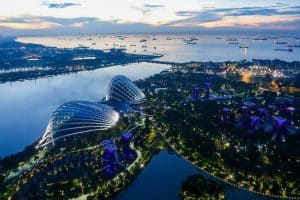 Why Singapore Belongs on Your Bucket List 24 Aerial view of Gardens by the Bay in Singapore at dusk, featuring illuminated glass conservatories and iconic Supertree structures near the waterfront with ships anchored offshore, showcasing the vibrant beauty of Singapore.