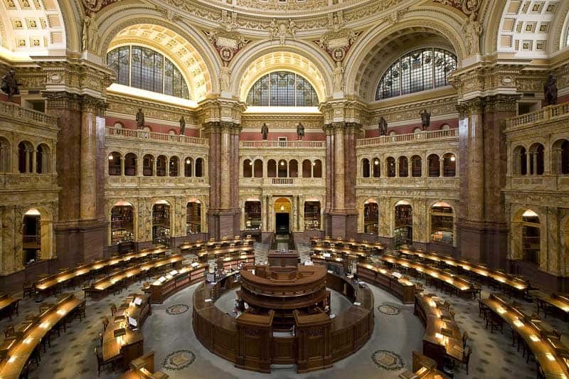 A large, ornate library reading room with a domed ceiling, arched windows, and circular rows of wooden desks and chairs surrounding a central desk area.