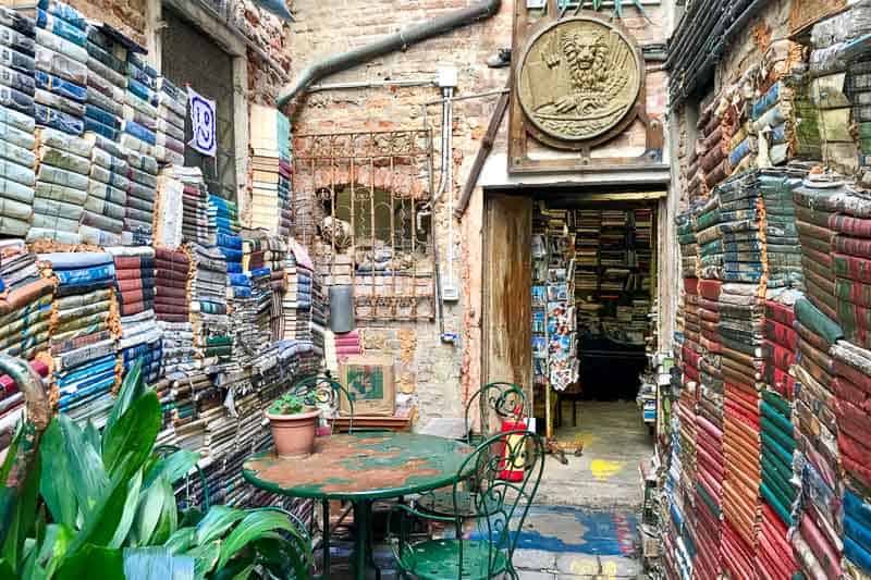 Stacks of old books line the walls of a narrow outdoor courtyard with a small round table, metal chairs, and potted plants; a bookshop interior is visible through an open doorway.